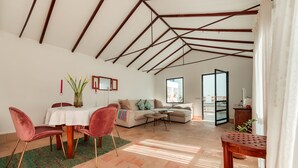 A spacious living area with a high ceiling, a dining table with chairs, a sofa, and a view through an open door.
