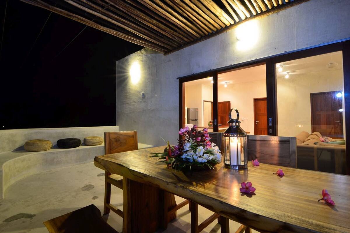 A wooden dining table with a flower arrangement and a lantern, set against a modern building with large windows.