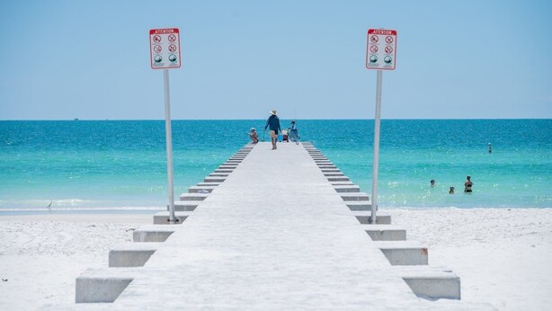 En brygga som leder till en strand med klart, blått vatten, där folk promenerar, och två uppmärksamhetsskyltar.