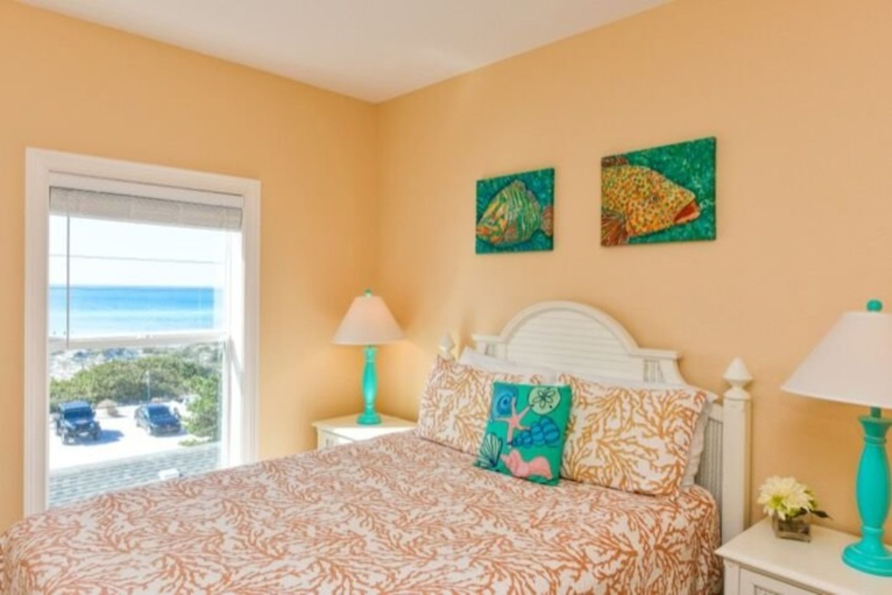A bedroom with a bed, two wall-mounted paintings, a window with a view, and a nightstand with a lamp.
