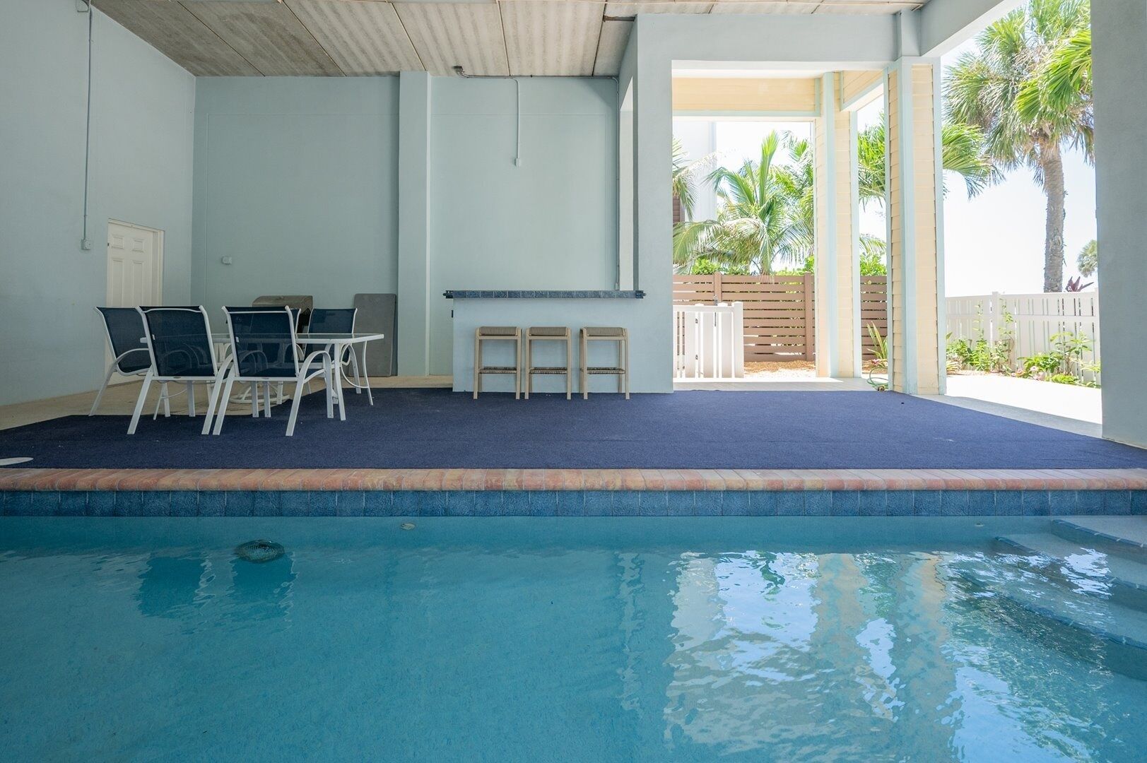 Ein Hallenbad mit blau ausgelegtem Teppichbereich, einem Tisch mit Stühlen und Blick auf einen umzäunten Außenbereich mit Palmen.