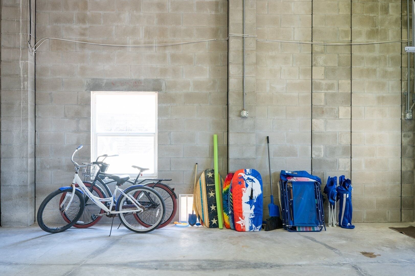 A room with a concrete floor, a window, a bicycle, and several bags and surfboards.