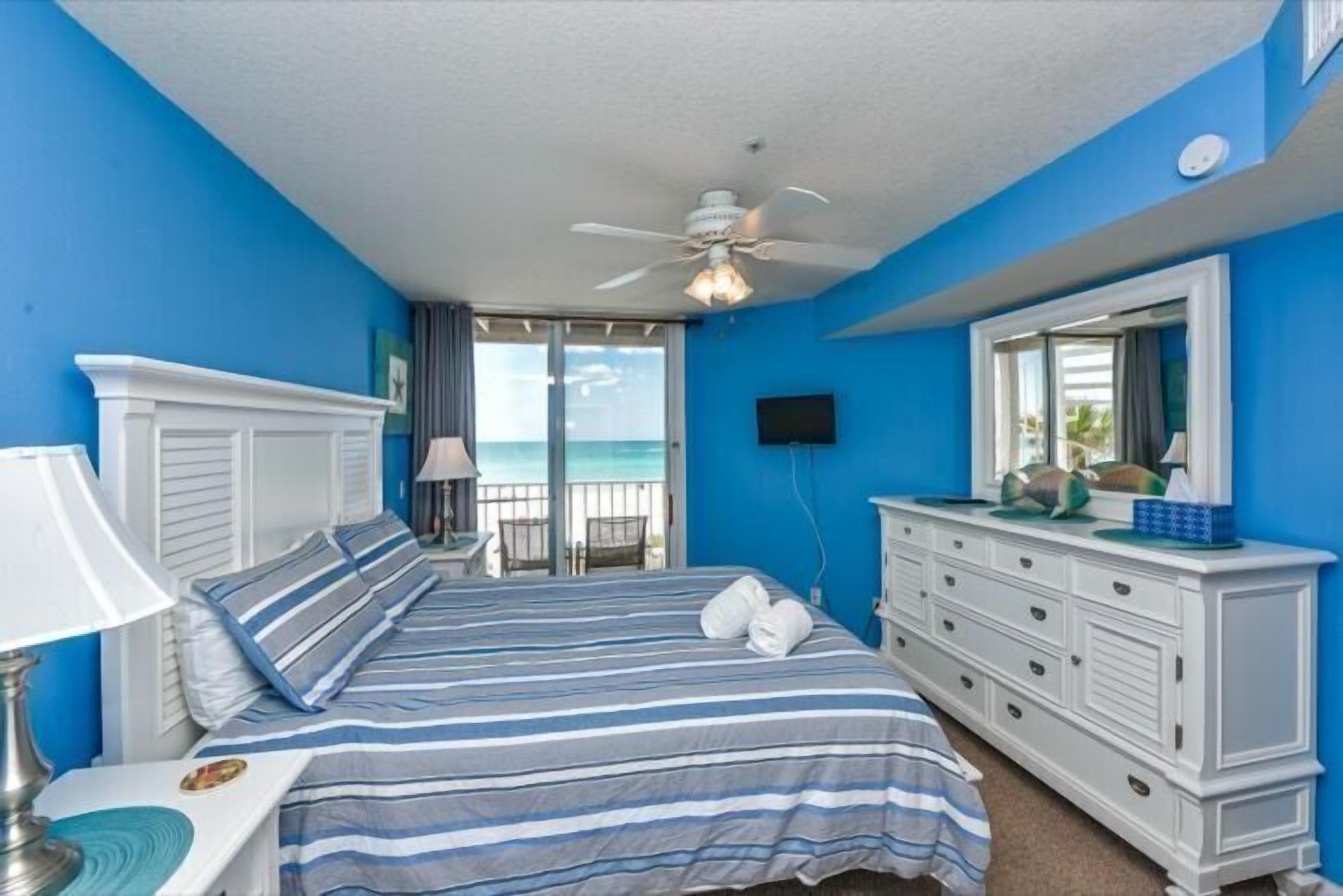 A hotel room with a large bed, a white dresser, a ceiling fan, and a view of the ocean.