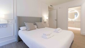 A modern hotel room with a large bed, two patterned pillows, a bedside table with a lamp, and a view of the bathroom. - Brunet II | The Rentals Collection (San Sebastián)