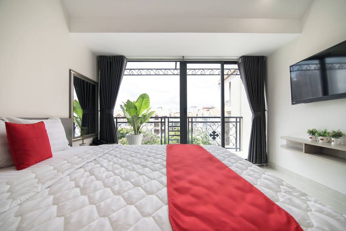 A hotel room with a large bed, a red throw, a TV, and a balcony with a view.