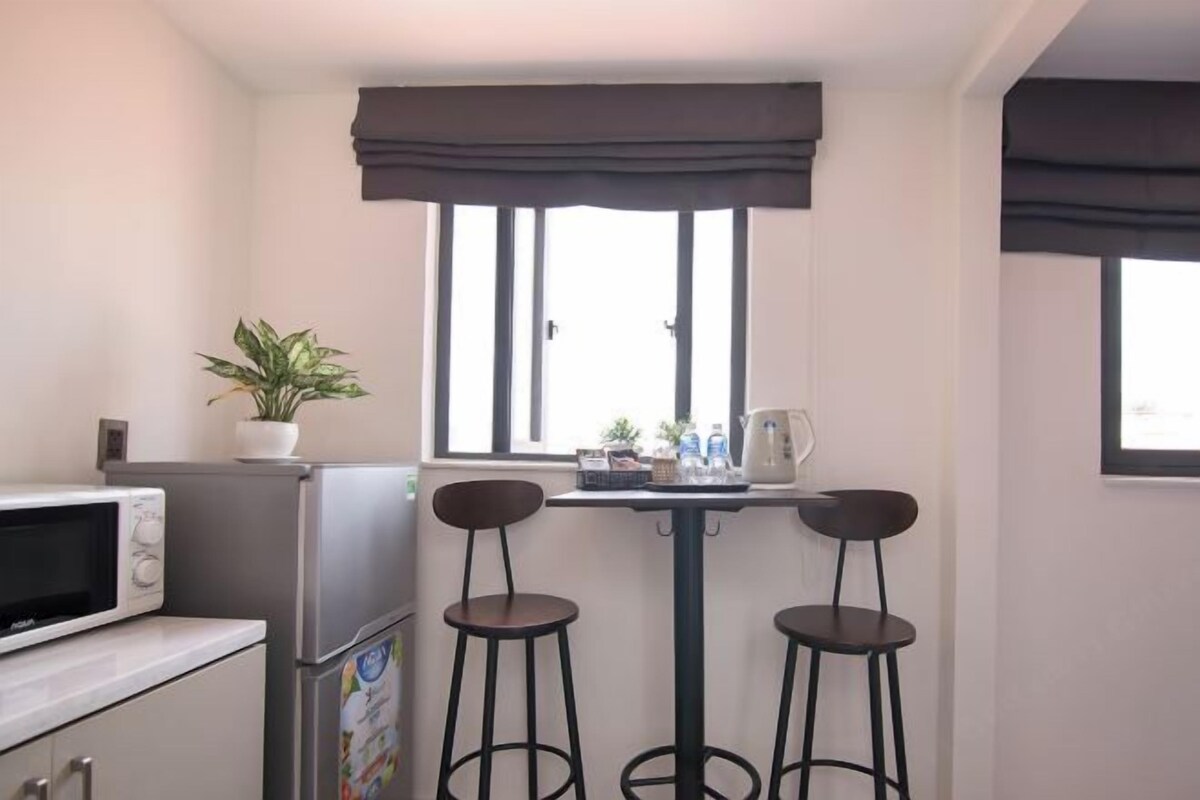 A compact kitchen with a microwave, refrigerator, and two bar stools. A window with a curtain is visible, and a potted plant is on the counter.