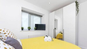 A modern bedroom with a yellow bedspread, a large window with blinds, a flat-screen TV, and a wardrobe with a mirror. - Little Corner of Bliss in BustlingLondon (London)