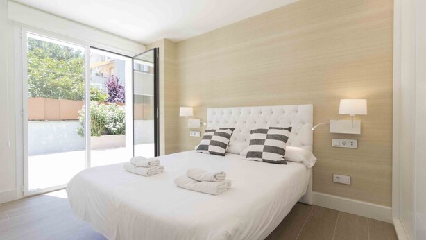 A hotel room with a large bed, two bedside lamps, a sliding glass door to a balcony, and a view of greenery outside. - Brunet I | The Rentals Collection (San Sebastián)