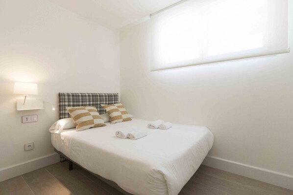 A neatly made bed with white bedding and striped pillows, a bedside lamp, and a window with blinds. - Brunet I | The Rentals Collection (San Sebastián)