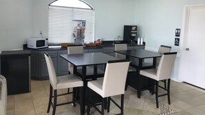 A compact kitchen with a microwave, coffee maker, and a beverage dispenser. There is a dining table with chairs and a white door with accessibility signs. - Crown Inn Humble (Humble)