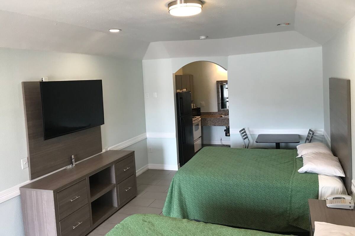 A bedroom with a TV, a desk, a bed, and a kitchenette.