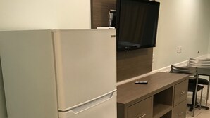 A compact kitchen with a white refrigerator, a wooden cabinet, a flat-screen TV mounted above the counter, and a small dining area with a chair. - Crown Inn Humble (Humble)