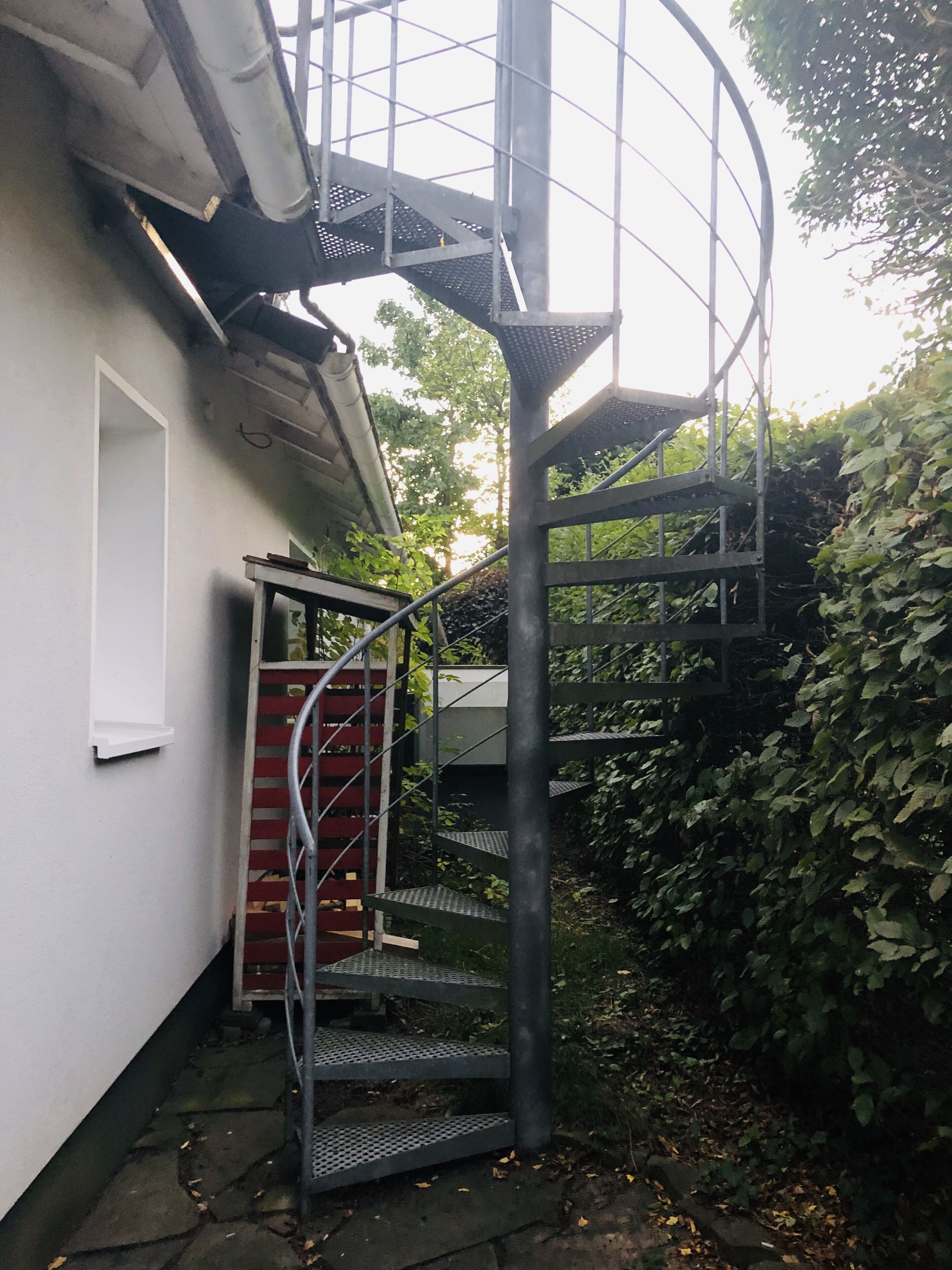 Eine Metall-Wendeltreppe führt zu einer zweiten Etage, neben einem weißen Gebäude mit einer roten Tür und einem kleinen Fenster.