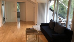 Un salon moderne avec un canapé noir, une table basse en verre et une vue sur un balcon aménagé avec du mobilier d’extérieur.