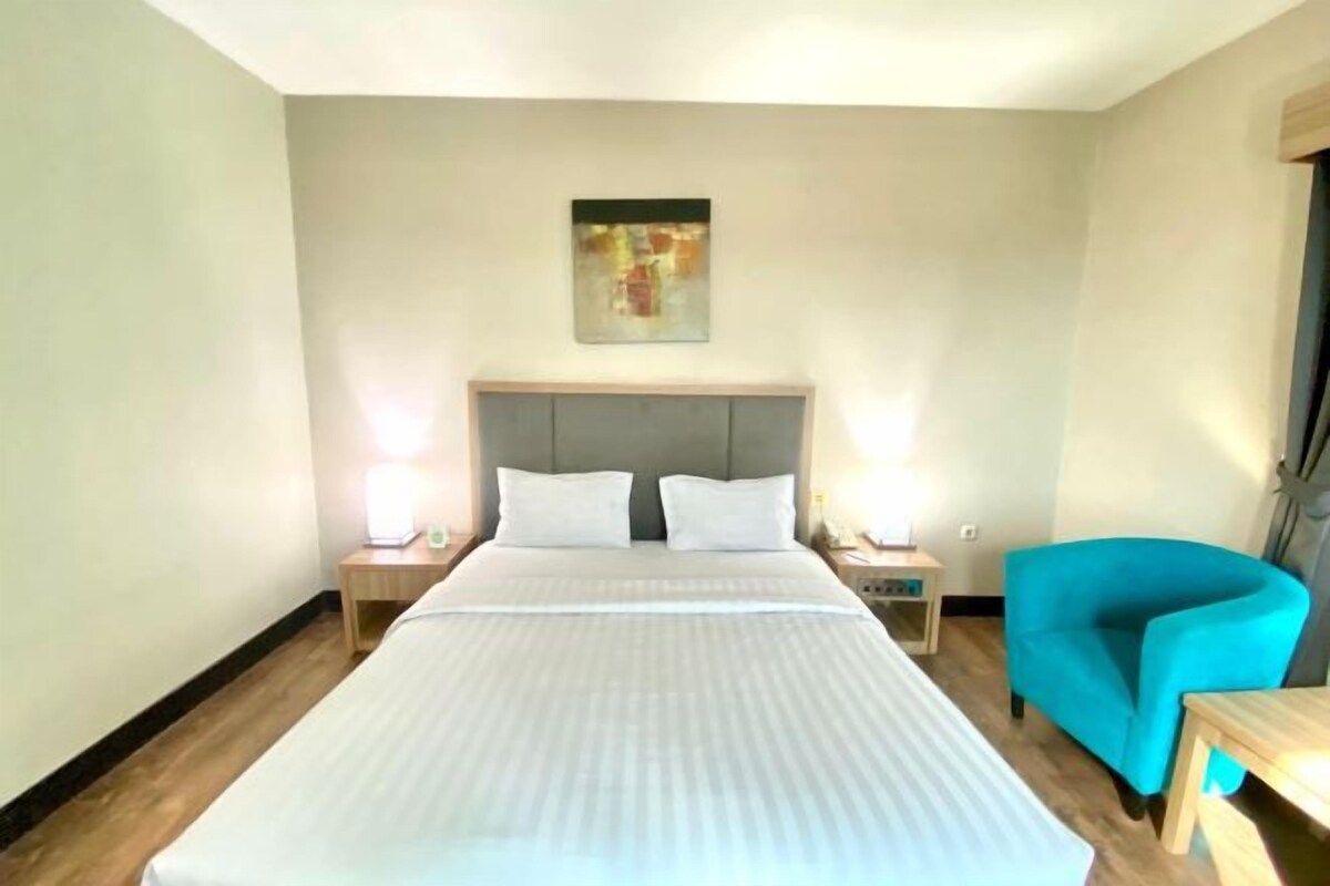 Une chambre d’hôtel avec un grand lit, deux tables de chevet, un fauteuil bleu et un tableau au mur.