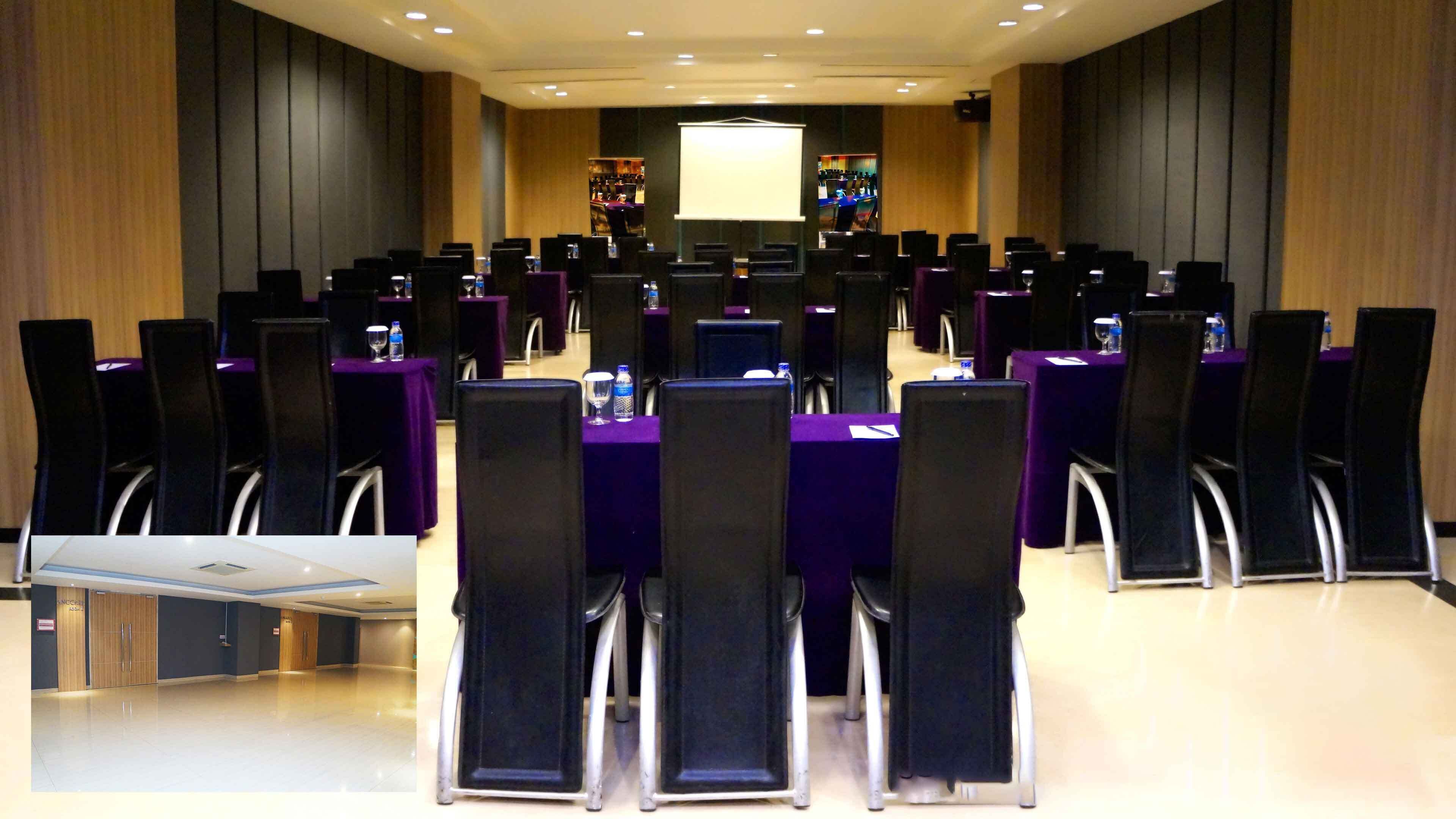 A conference room with rows of tables and chairs, a large screen at the front, and a bar area in the background.