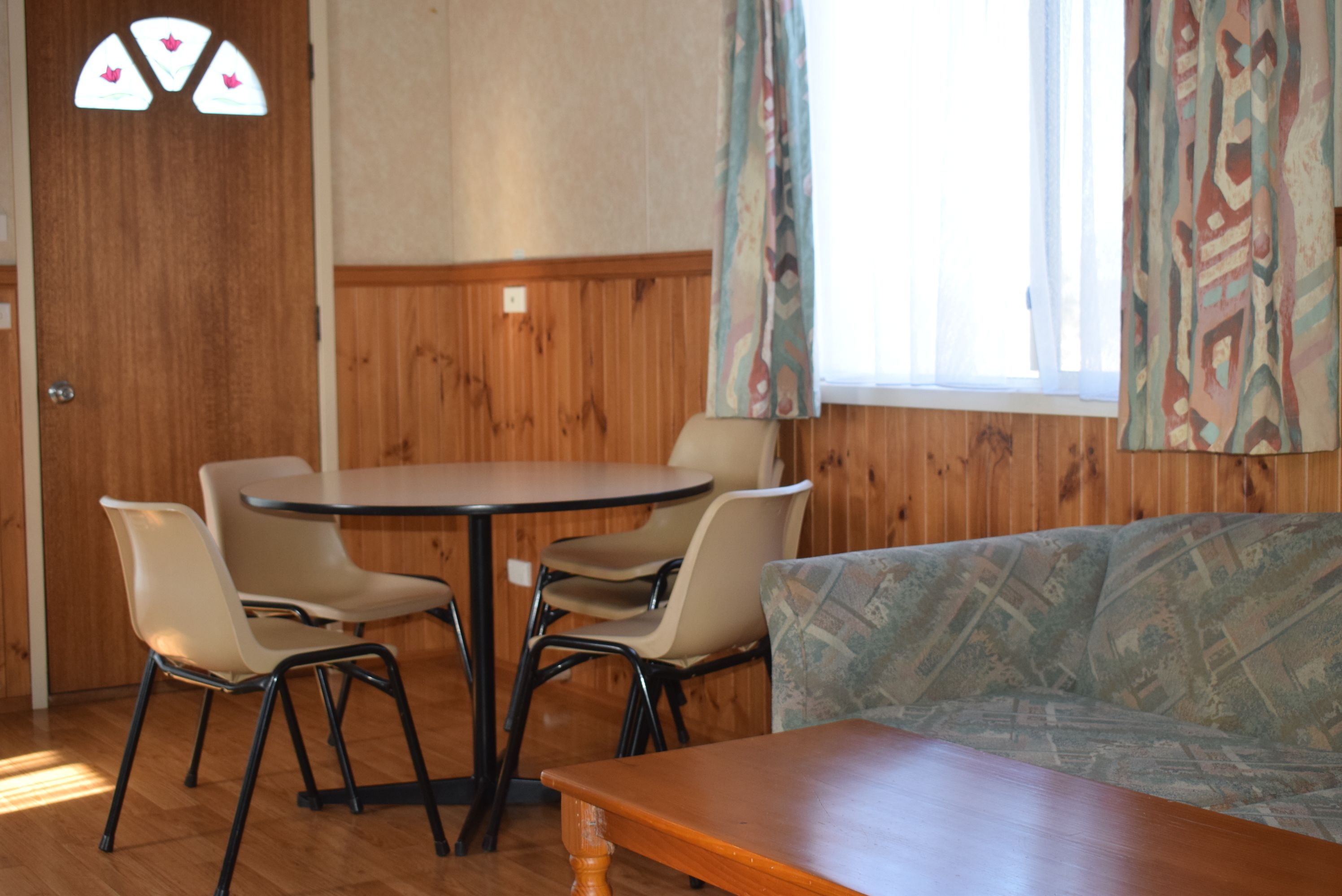 A room with a wooden paneled wall, a round table with four chairs, a sofa, and a window with patterned curtains.