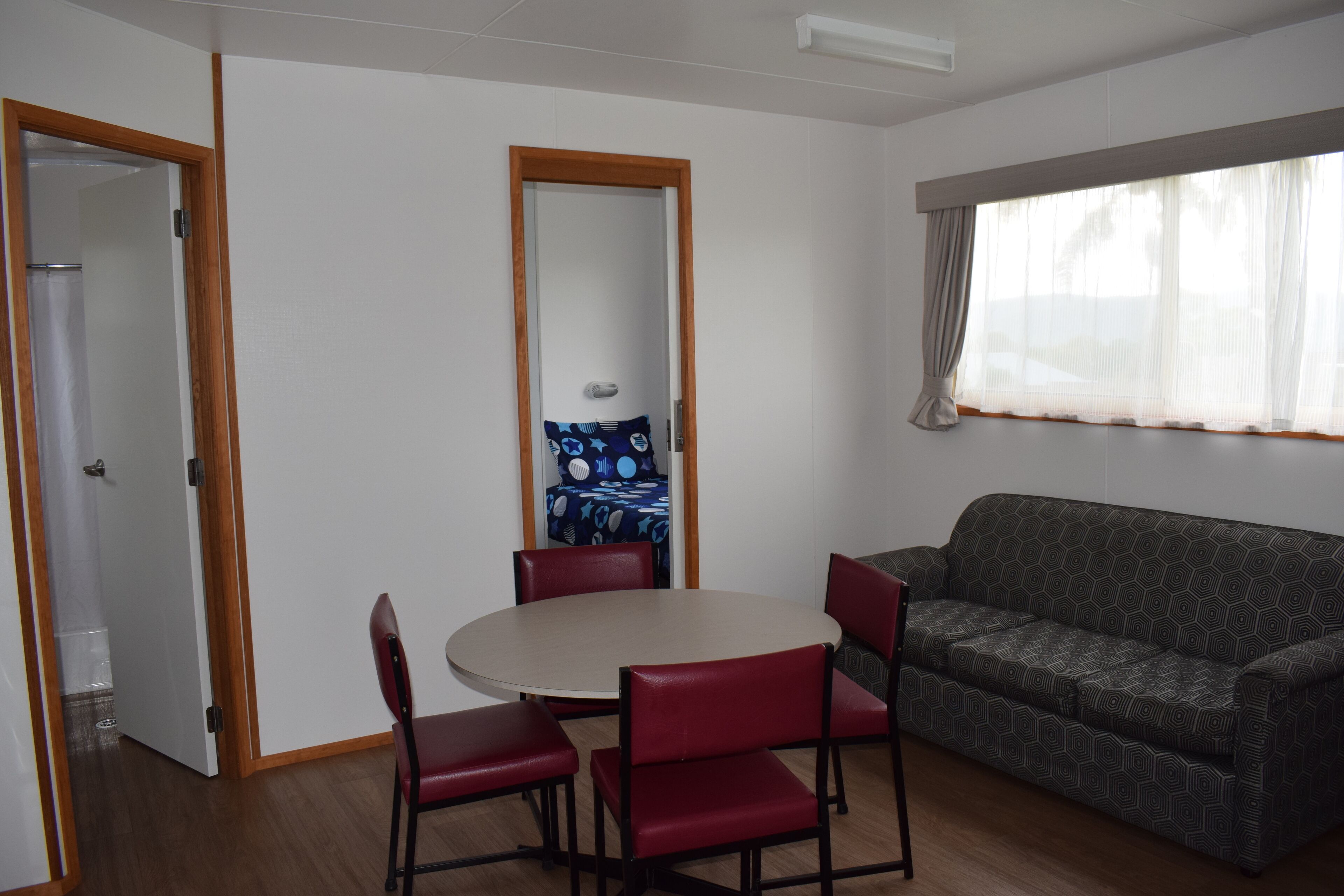 Una habitación con una mesa redonda, cuatro sillas rojas, un sofá gris con estampado y un espejo grande, además de una ventana con cortinas.
