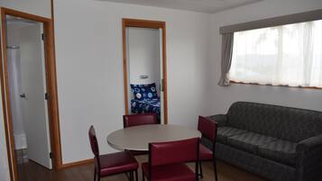 Una habitación con una mesa redonda, cuatro sillas rojas, un sofá gris con estampado y un espejo grande, además de una ventana con cortinas.