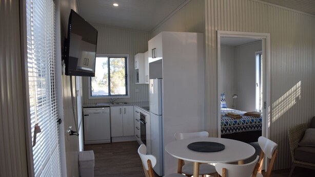 A compact kitchen with white cabinets, a built-in oven, and a refrigerator. There is a dining area with a round table and white chairs. - Bega Caravan Park (Bega)