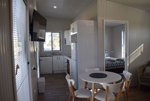 A compact kitchen with white cabinets, a built-in oven, and a refrigerator. There is a dining area with a round table and white chairs. - Bega Caravan Park (Bega)
