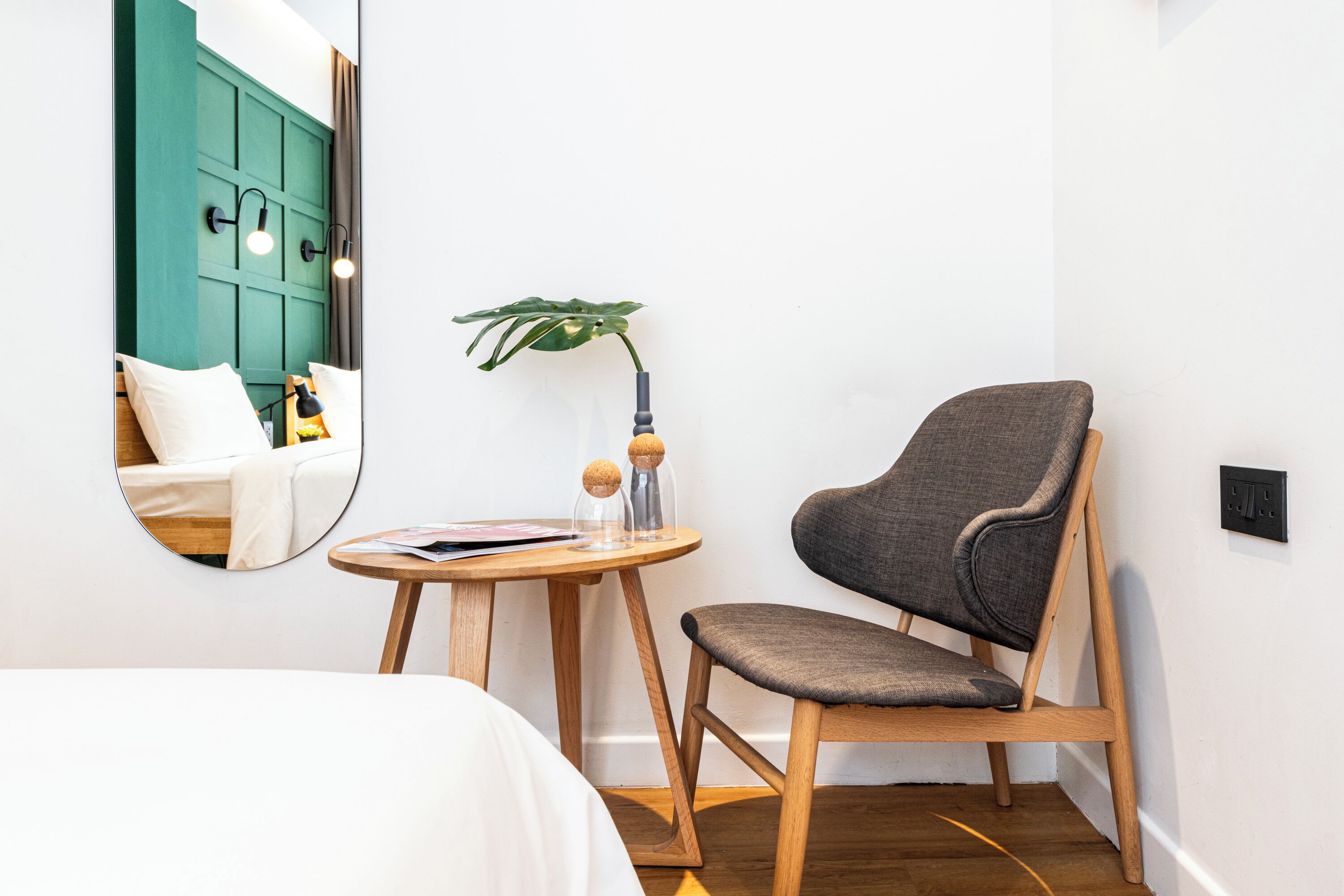 A compact hotel room with a bed, a round wooden table, a chair, a mirror, and a potted plant.