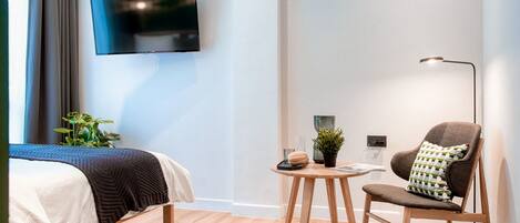 A modern hotel room with a bed, a chair, a small table with a plant, and a TV mounted on the wall.