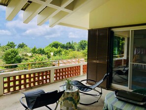 A balcony with a view of greenery, a chair, a small table with a plant, and a sliding glass door.