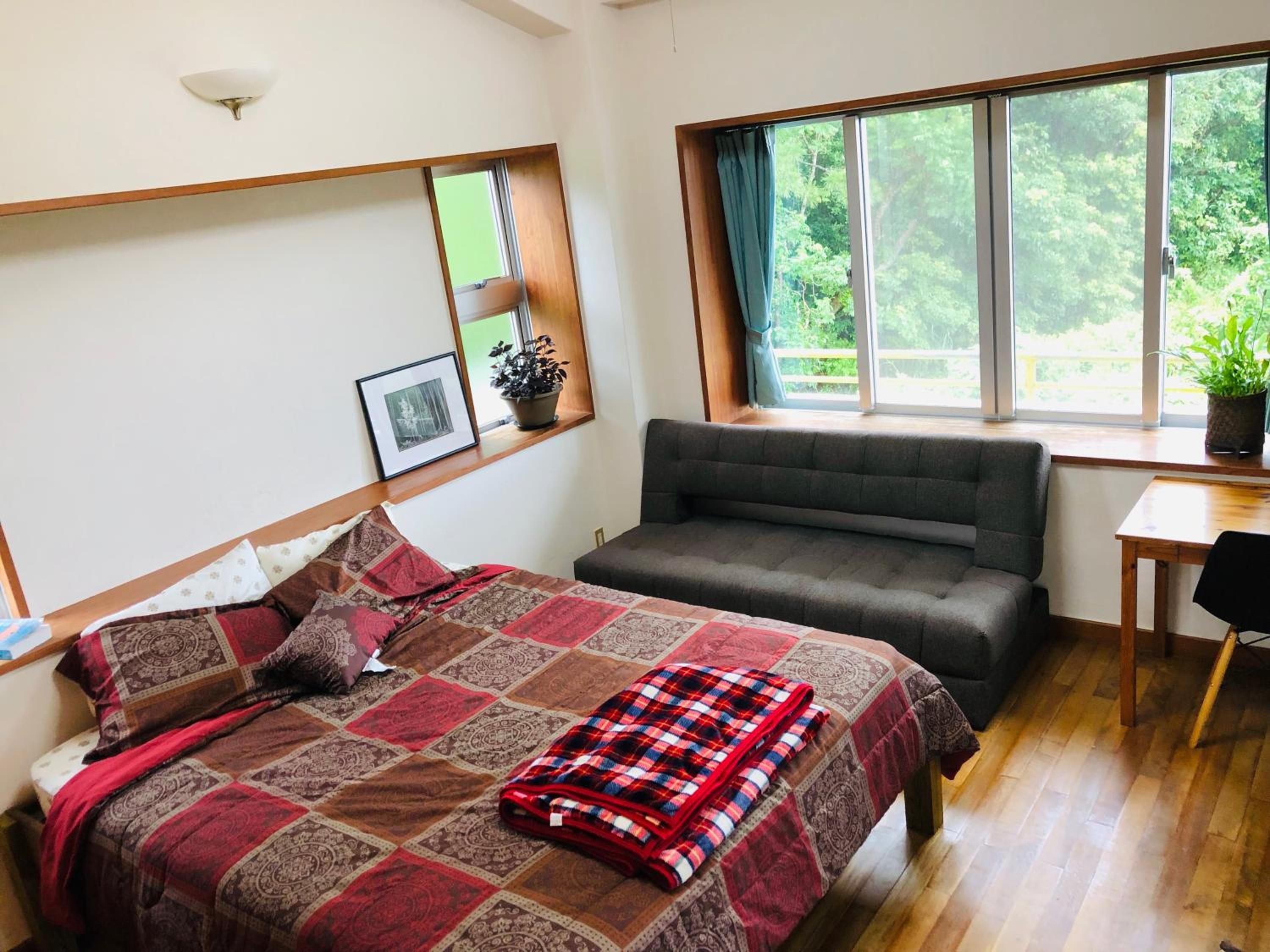 A bedroom with a bed, a sofa, a window with curtains, a small table, and a potted plant.