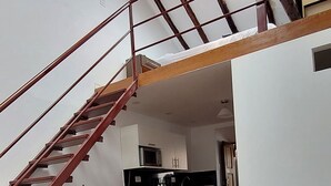 A loft-style room with a wooden staircase, a dining area with stools, a sofa, and a kitchen in the background. - Hospedaje Laura (Cusco)