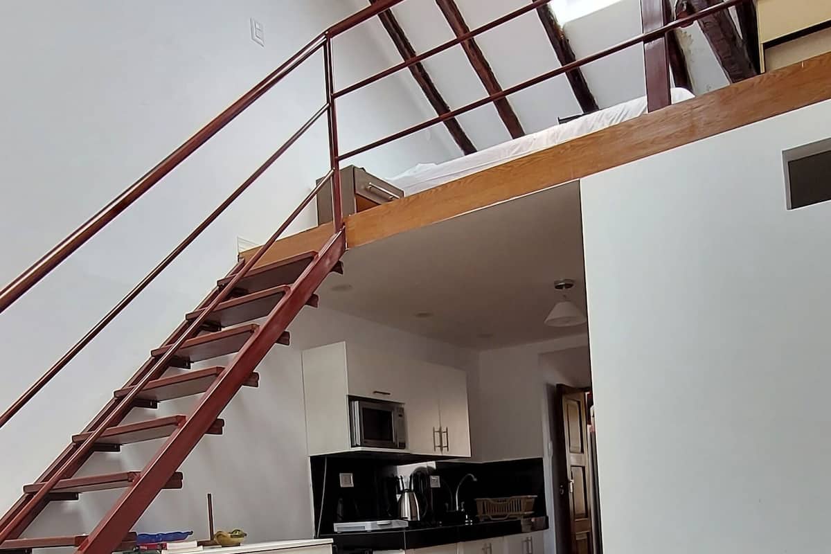 A loft-style room with a wooden staircase, a dining area with stools, a sofa, and a kitchen in the background.