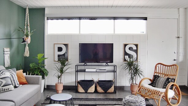 A living room with a grey sofa, a wicker chair, a TV on a stand, and two potted plants. - Mid-Century Modern Bohemian Bungalow w/ Pool (Palm Springs)