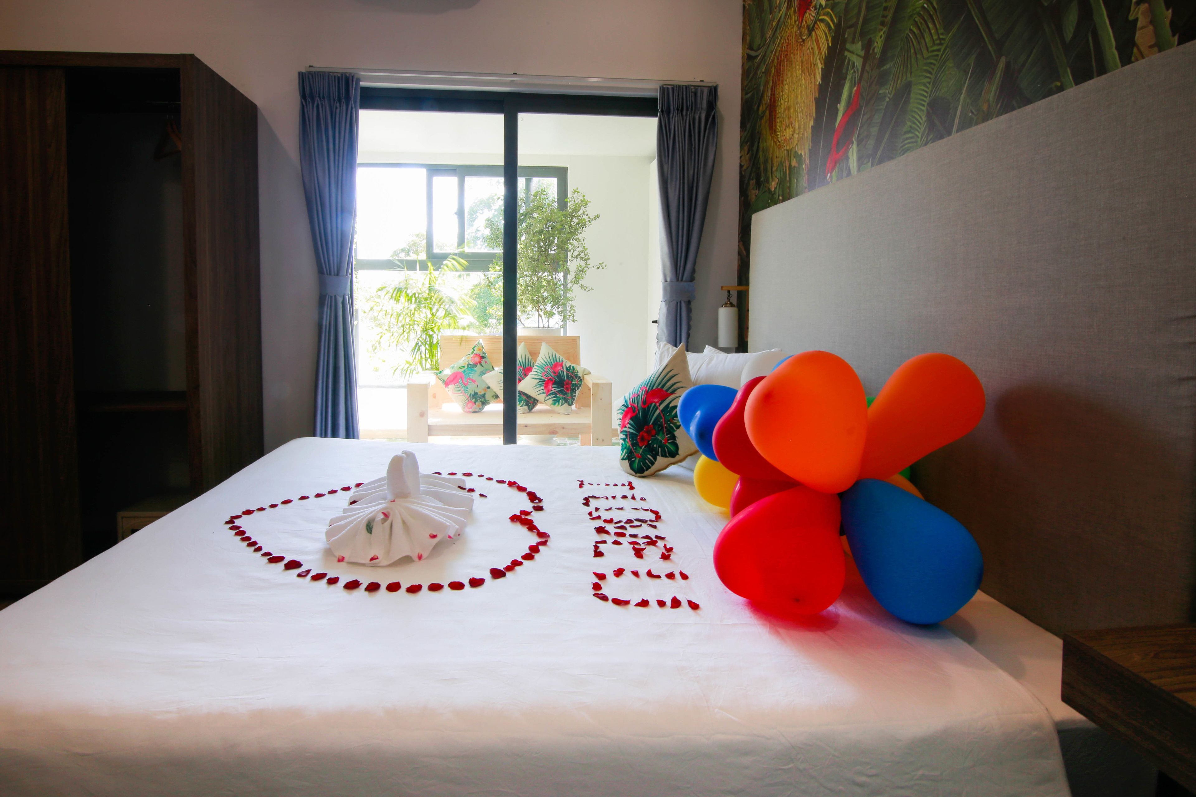 A neatly made bed with a white sheet, a towel, and rose petals arranged in a heart shape. There are colorful balloons on the bed.