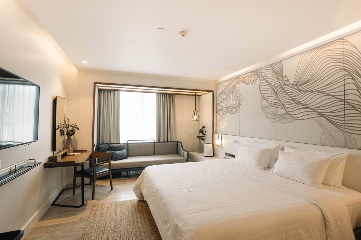 Una habitación de hotel moderna con una cama grande, un escritorio con silla, un sofá y un ventanal con cortinas.