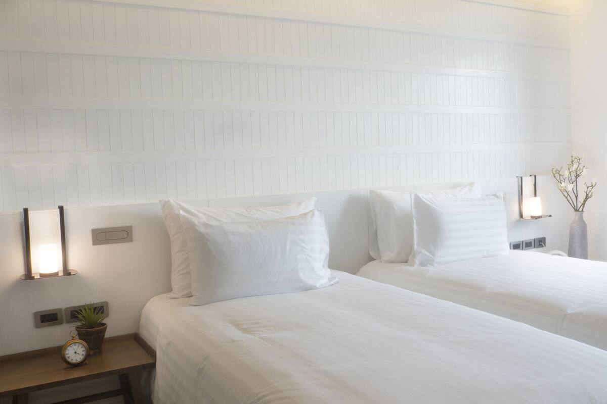 Habitación de hotel con dos camas, una mesita de noche con un reloj y una planta, y un aplique de pared.
