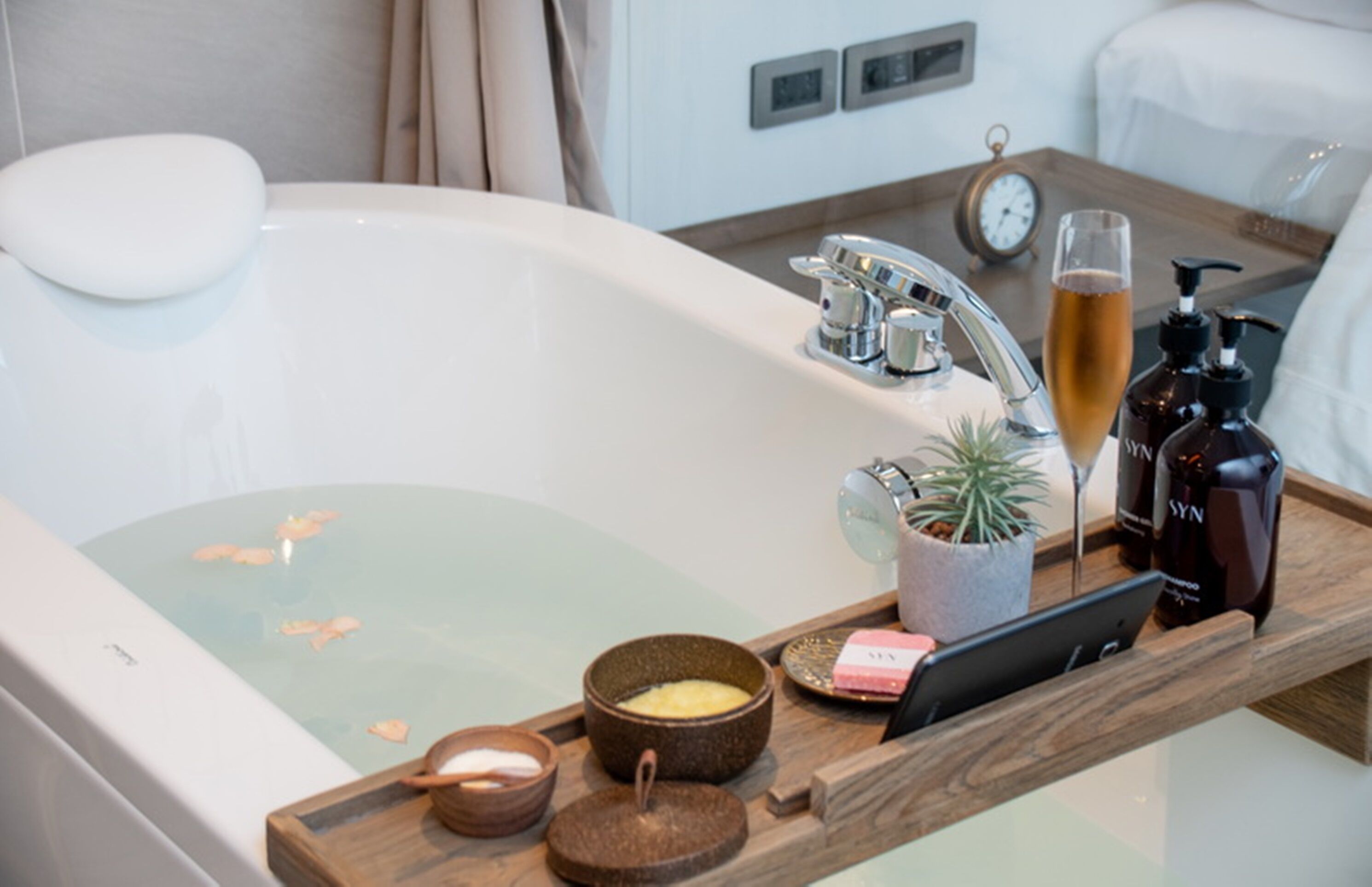 A bathroom with a bathtub, a wooden tray with toiletries, a clock, and a plant.