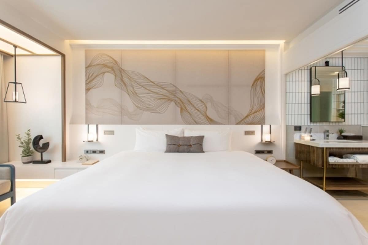 Una habitación de hotel moderna con una cama grande, una mesita de noche con lámpara, un espejo y un baño con lavabo y tocador.