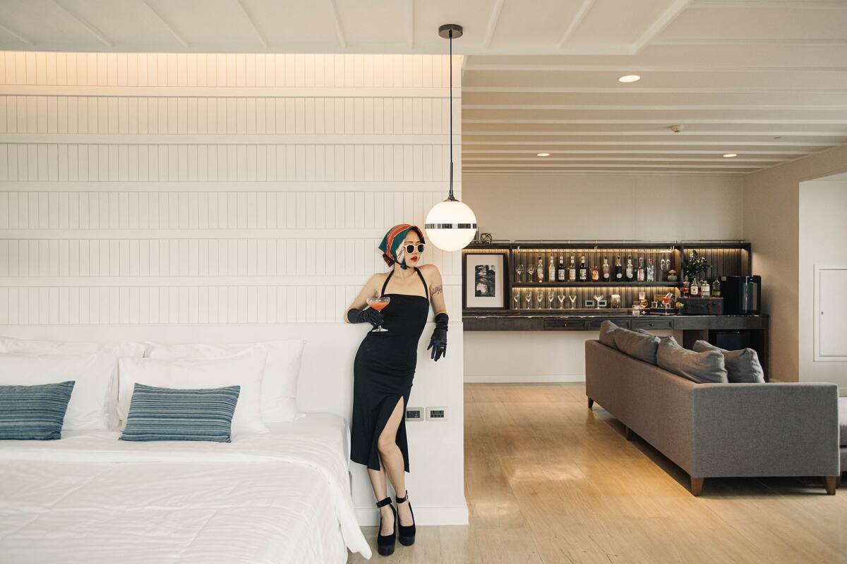 Una habitación de hotel moderna con una cama, un sofá gris y una zona de bar al fondo.