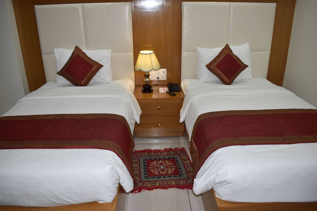 Habitación de hotel con dos camas, cada una con colcha roja y blanca, cabecero de madera, mesita de noche con lámpara y alfombra roja estampada.