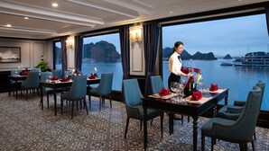A dining area with tables set for two, a waiter attending to a table, and a view of a body of water with a boat in the distance. - Doris Cruise (Tuan Chau)