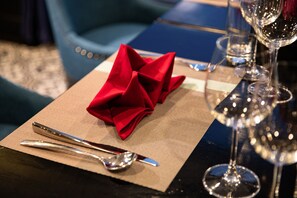 A table set with a folded red napkin, a spoon, and a fork on a placemat, with wine glasses in the background. - Doris Cruise (Ha Long)