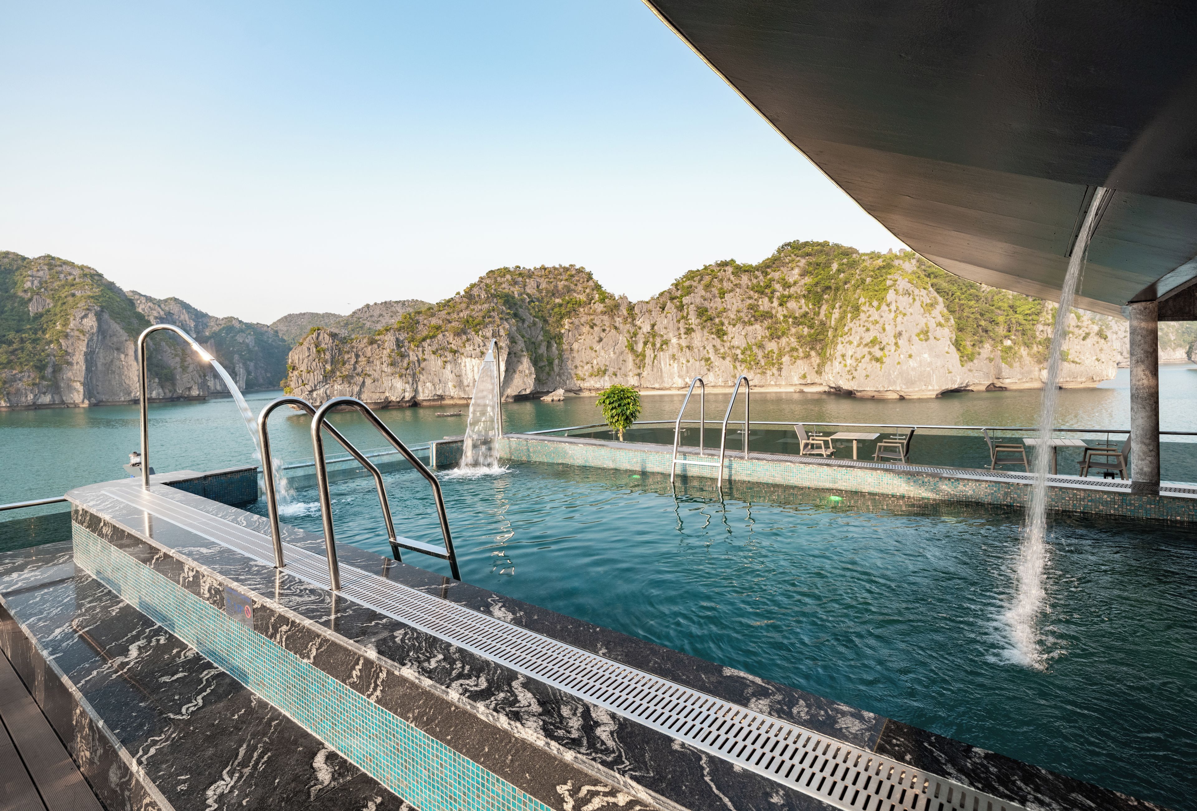 Une piscine à débordement offrant une vue sur un paysage montagneux, agrémentée d’un élément aquatique et d’un espace abrité avec des sièges.