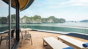 A deck with wooden flooring, lounge chairs, and a glass railing overlooking a body of water with rocky islands.