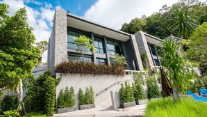 Um edifício moderno com pilares de pedra, grandes janelas e um jardim ajardinado com diversas plantas e uma palmeira.