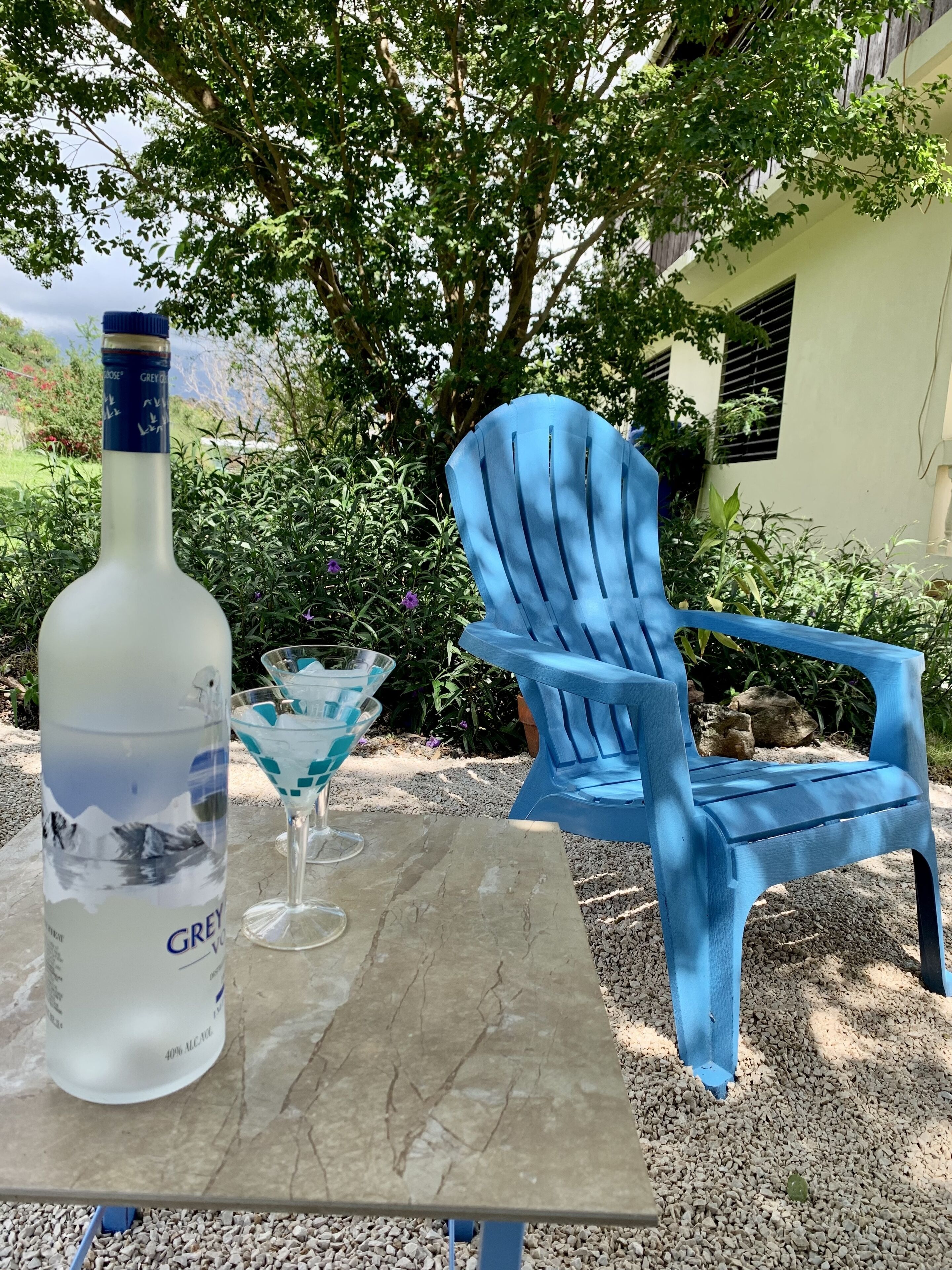 Une table avec une bouteille de vodka Grey Goose, un verre à martini et une chaise Adirondack bleue, le tout installé dans un décor extérieur agrémenté de verdure et avec un bâtiment en arrière-plan.
