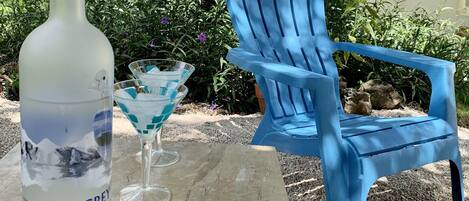 Une table avec une bouteille de vodka Grey Goose, un verre à martini et une chaise Adirondack bleue, le tout installé dans un décor extérieur agrémenté de verdure et avec un bâtiment en arrière-plan.