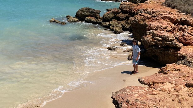 Une plage rocheuse avec une eau claire, et une personne debout sur le sable.