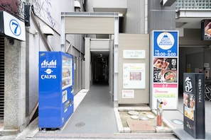 Une ruelle étroite où l’on aperçoit un distributeur automatique bleu portant l’inscription « Asahi Soft Drinks CALPIS » et un magasin fermé, sur lequel est affiché un panneau indiquant « Dioré Crest ».