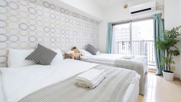 A hotel room with two beds, a potted plant, and a view of the city through the window. - Shinsaibash East Nana Apartment · Junan (Osaka)