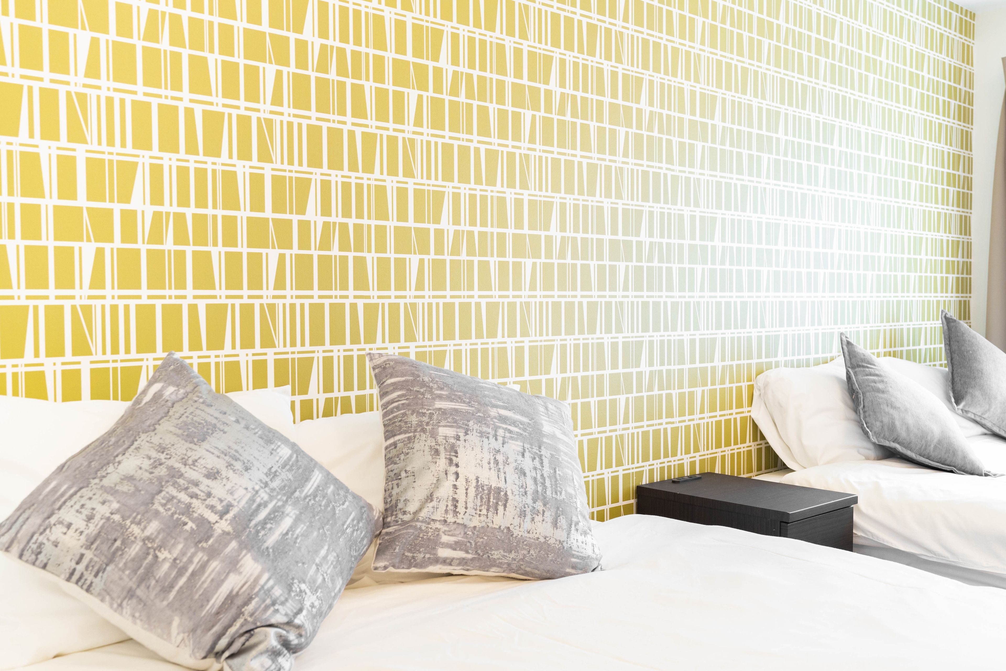 A hotel room with a yellow patterned wall, a bed with white bedding, and two gray pillows.